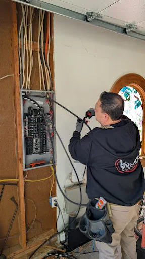 A technician working beside an open electrical panel and exposed wall wiring in a garage